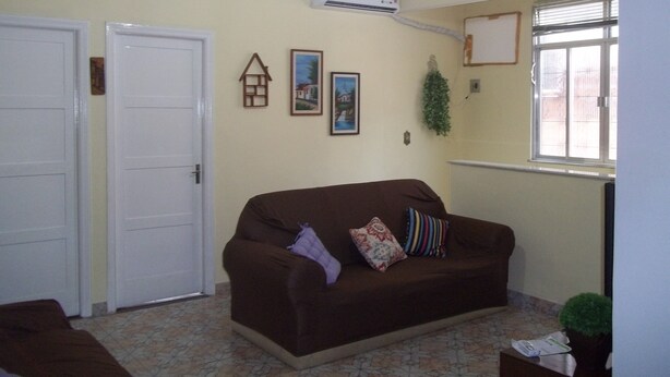 Ein Wohnzimmer mit braunem Sofa, einer weißen Tür, einer Klimaanlage, einem Fenster mit Jalousien und einer kleinen Pflanze.