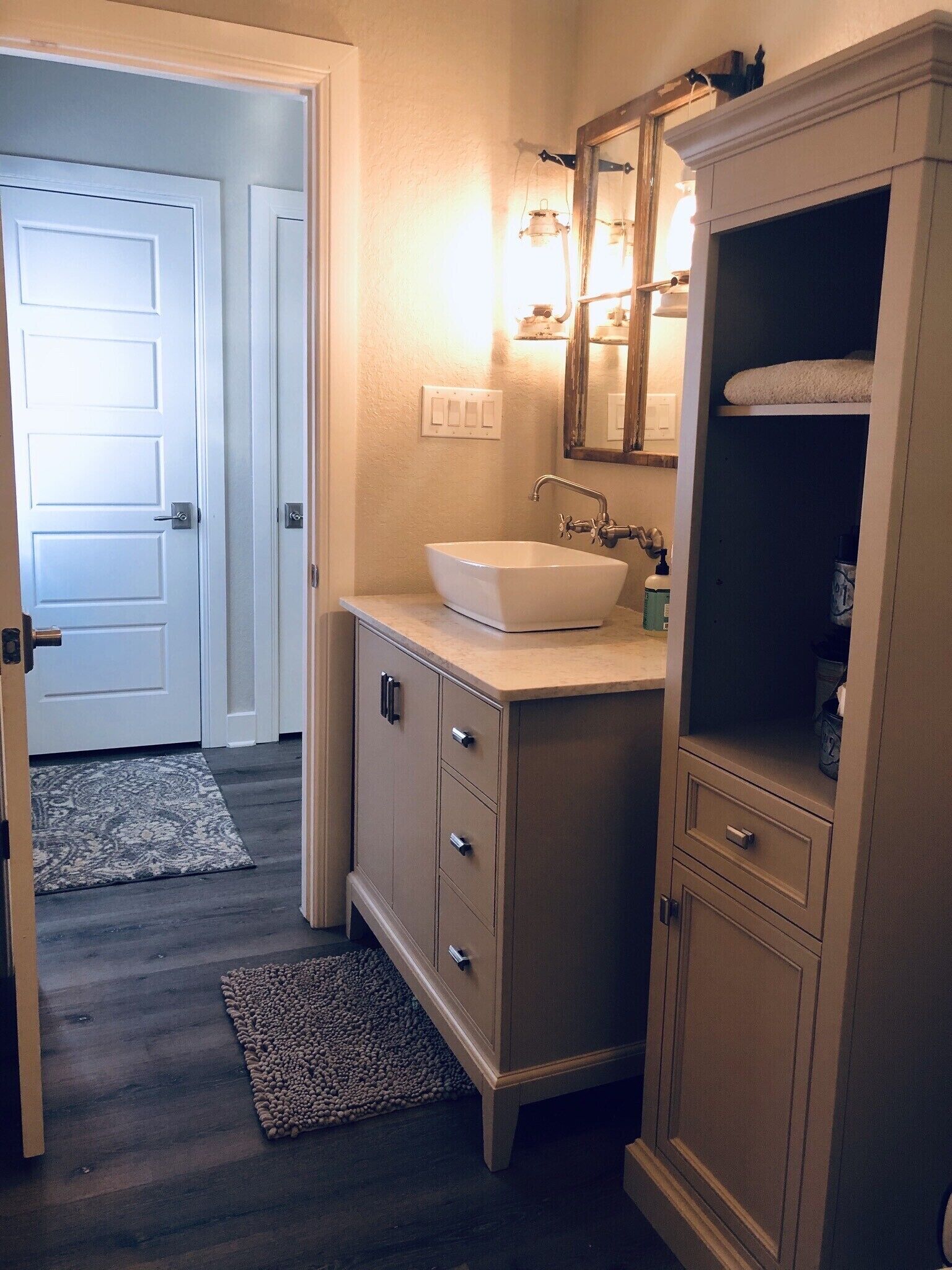 Une salle de bain avec un lavabo blanc, un miroir au-dessus et un meuble avec des tiroirs. Il y a un tapis sur le sol et une porte au fond.