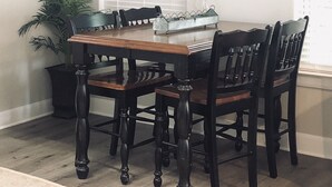 A dining area with a wooden table and four chairs, a potted plant, and a wall-mounted sign with the word "gather".