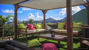 An outdoor seating area with wooden furniture, cushions, and a view of mountains and the sea. - Located on the famous island of St. Barts, Villa Eugenie is a charming house.  (Saint Barthélemy)