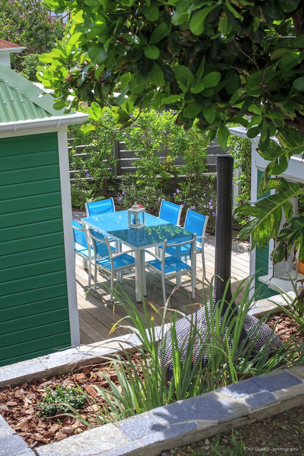 Une terrasse avec une table et des chaises, entourée de verdure et d’une clôture verte.
