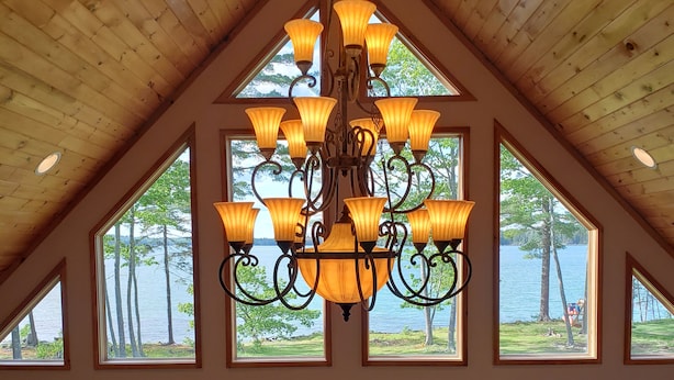 Une pièce spacieuse avec un haut plafond en bois, un grand lustre, de nombreuses fenêtres donnant sur un lac et une cheminée en pierre.