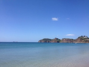 A coastal scene with clear blue water, a sandy beach, and a rocky hill with buildings. - Spectacular Condominio View of Flamingo Beach and Potrero Bay   (Playa Flamingo)