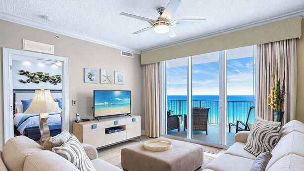 Una sala de estar con televisor, sofá, reposapiés, ventilador de techo y un balcón con vista al océano.