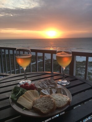 Une table avec deux verres de vin, une assiette de fromage et du pain, et une vue sur le coucher de soleil sur l’océan.