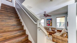 Un escalier en bois mène à un salon équipé d’un ventilateur de plafond, d’un canapé et d’une fenêtre avec des stores.
