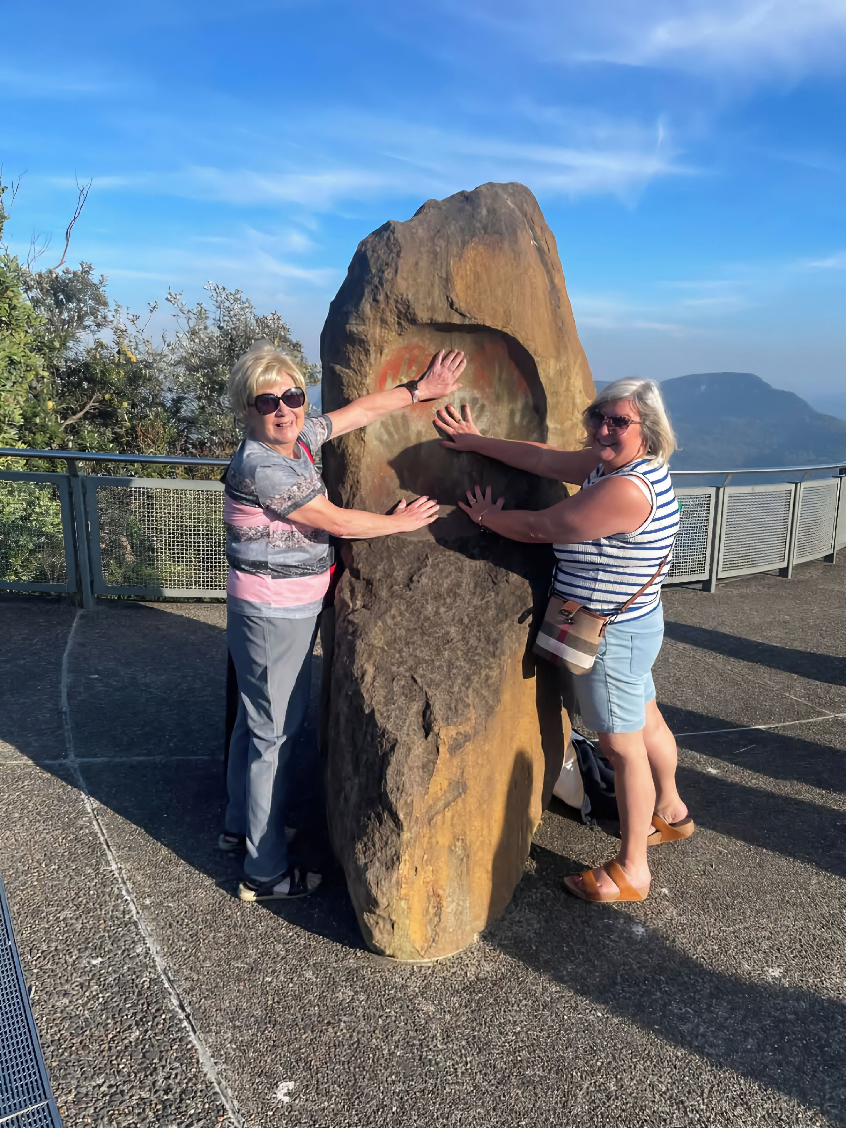 Two people touching a large rock with a scenic view in the background.