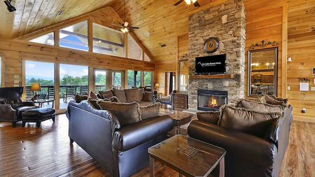 A spacious living room with leather sofas, a stone fireplace, and large windows offering a view of the outdoors. - Gatliburg Mansion | Best Views In The Smokies | 14 Full Suites (Gatlinburg)