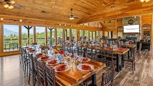 A spacious dining area with wooden tables and chairs, large windows offering views of the outdoors, and a stone fireplace. - Gatliburg Mansion | Best Views In The Smokies | 14 Full Suites (Gatlinburg)
