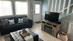 A living room with a black sofa, a coffee table, a TV on a wooden stand, and a large window with blinds. - Beautiful Gulf Front Complex,White Sandy Beaches, private pool, Paradise Awaits! (Navarre)