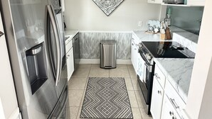 A modern kitchen with a stainless steel refrigerator, microwave, and oven. There is a trash can, a patterned rug, and a decorative wall piece. - Beautiful Gulf Front Complex,White Sandy Beaches, private pool, Paradise Awaits! (Navarre)