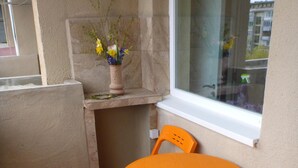 Un petit balcon avec une table ronde orange et des chaises assorties, une plante en pot posée sur une étagère, et une vue sur les bâtiments à travers une fenêtre.