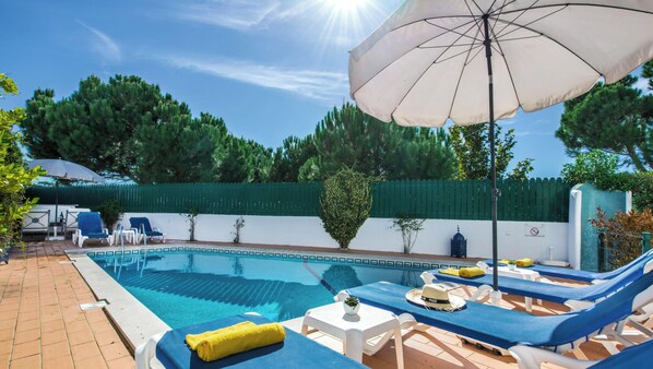 Zona de piscina con sillones, una mesa y una sombrilla.