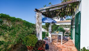 A patio with a table and chairs, surrounded by potted plants and a pergola with hanging vines.