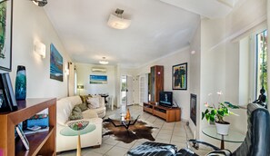 A modern living room with a sofa, coffee table, and TV. There is a glass table with a plant, and a leather chair.