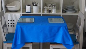 A dining area with a blue tablecloth, white chairs, and a white shelving unit with various kitchenware.