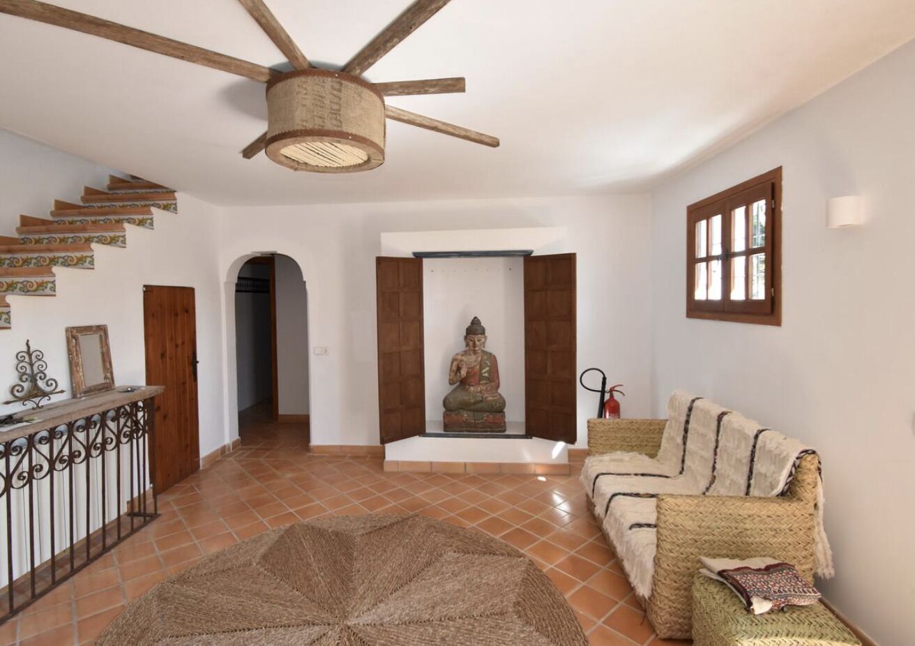 A spacious living area with a high ceiling fan, a Buddha statue, a woven sofa, and a staircase with decorative tiles.