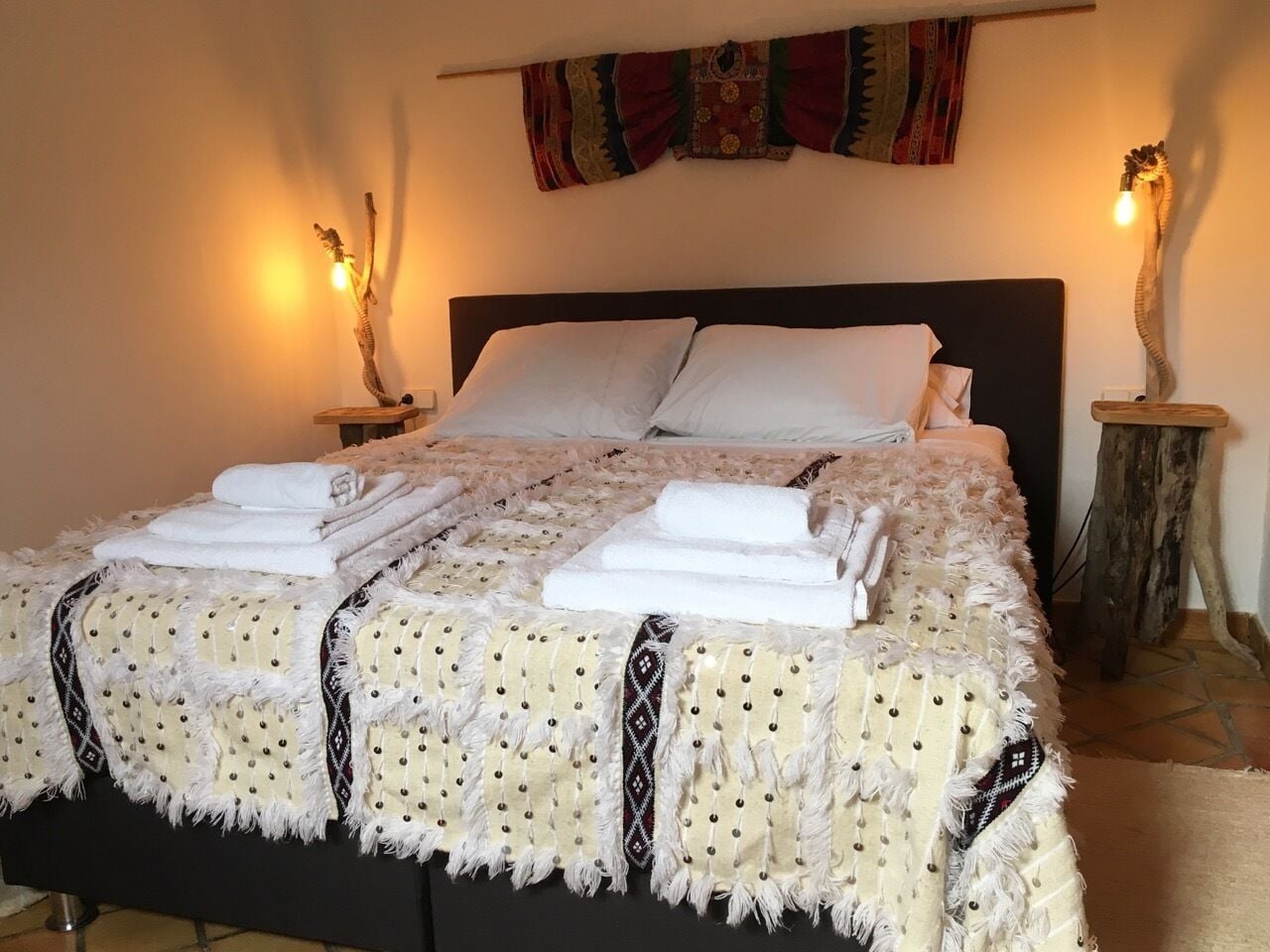 Een netjes opgemaakt bed met witte lakens en een bedekking met een patroon, met aan weerszijden wandlampen en een decoratief wandtapijt erboven.