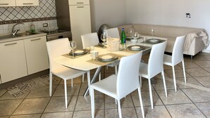 Un comedor con una mesa dispuesta para cuatro, sillas blancas, un sofá y una cocina al fondo.