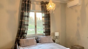 A bedroom with a bed, curtains, a chandelier, an air conditioner, and a bedside table with a lamp.