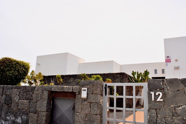 Un muro de piedra con una puerta y un cartel con el número 12, que da paso a un edificio moderno con tejado plano y fachada blanca.