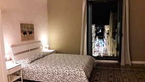 Une chambre d’hôtel avec un lit, une table de chevet, une lampe et une vue sur le paysage urbain, visible à travers une porte ouverte.