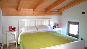 Une chambre avec un plafond en bois, un lit recouvert de draps blancs, une couvre-lit verte, deux tables de chevet avec des lampes rouges et une fenêtre donnant sur un beau paysage.