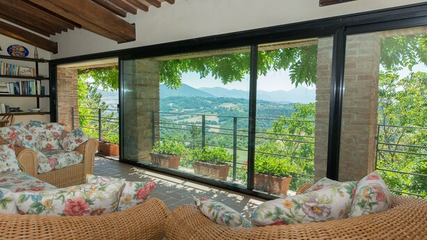 A room with wicker furniture, a bookshelf, and a large window with a view of a green landscape. - CHARMING FARMHOUSE IN THE MARCHIGIAN HILLS (Fabriano)