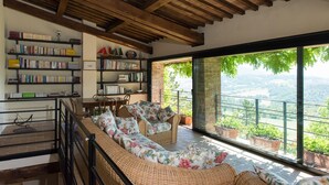 A room with a large window overlooking a scenic landscape, a bookshelf filled with books, and a wicker sofa with floral cushions. - CHARMING FARMHOUSE IN THE MARCHIGIAN HILLS (Fabriano)