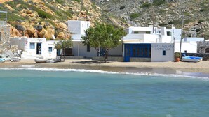 Ein Küstenhaus mit blauer Tür, weißen Wänden und einem Baum davor. Boote liegen in der Nähe.
