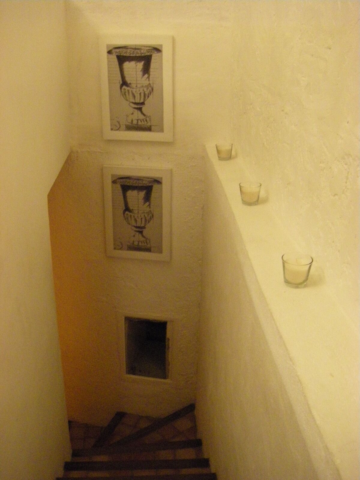 Een smalle trap met twee ingelijste foto’s aan de muur, een klein raam en enkele kaarsen op een plank.