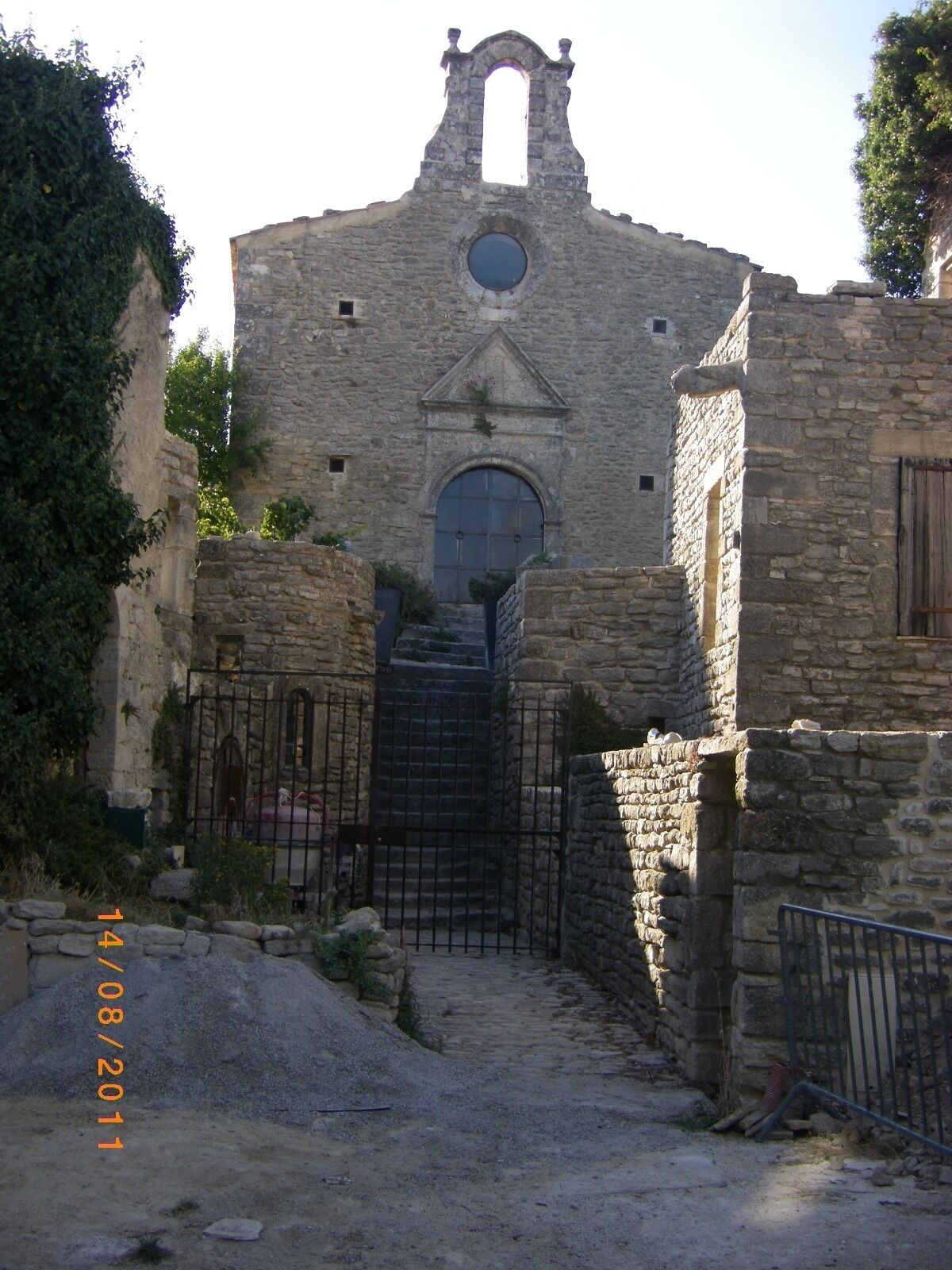 Un edifici de pedra antic amb una porta d'arc central, una finestra circular a la part superior de la porta i un petit balcó amb barana.