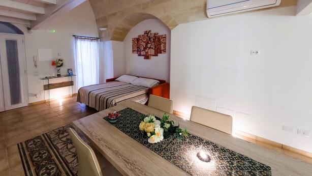 A hotel room with a wooden dining table, a bed with striped bedding, a decorative wall piece, and an air conditioning unit. - Cozy and comfortable apartment in Matera (Matera)