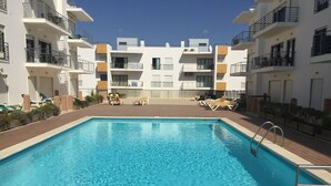 Ein rechteckiger Swimmingpool mit klarem, blauem Wasser, umgeben von einer gefliesten Liegefläche. Es befinden sich Liegestühle und ein kleiner Tisch am Pool. Der Pool grenzt an ein Gebäude mit Balkonen und Fenstern.