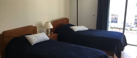 A room with two beds, a nightstand with a lamp, a wall-mounted light, and a view of the outside through a window.