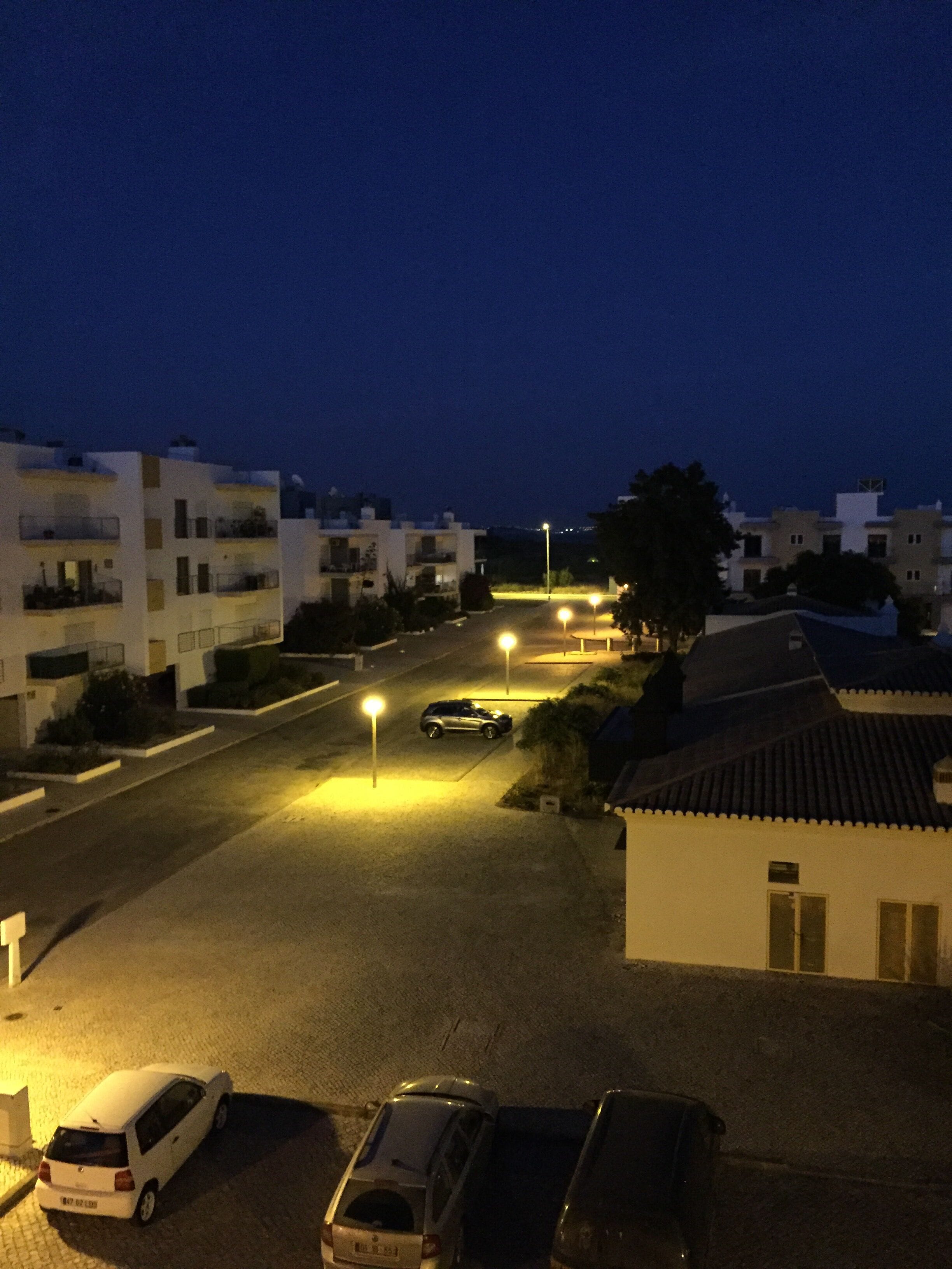 Un quartier résidentiel de nuit, avec des lampadaires, des voitures garées et des immeubles dotés de balcons.