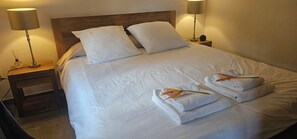 Un lit bien fait, avec du linge de lit blanc, deux oreillers et deux serviettes pliées, chacune ornée d’une fleur. Deux lampes de chevet sont posées sur des tables de nuit en bois.