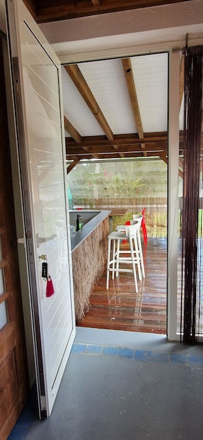 An open sliding door leading to a covered outdoor area with a wooden ceiling, a bar counter, and white chairs. - F1 Bungalow in beautiful Art Deco property (Petit Bourg)