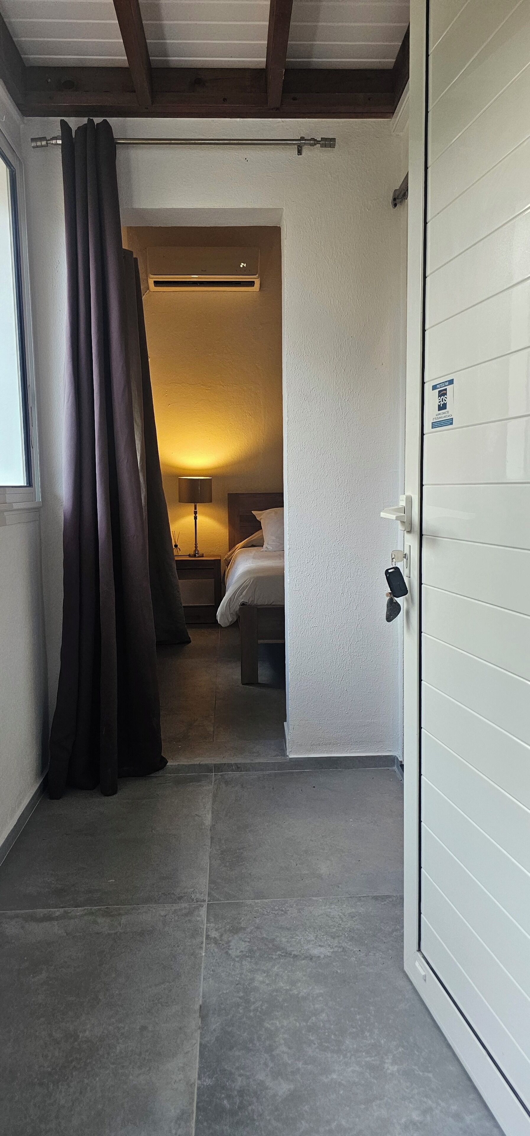 A hotel room with a bed, a nightstand, a lamp, a door, and a view of another room.