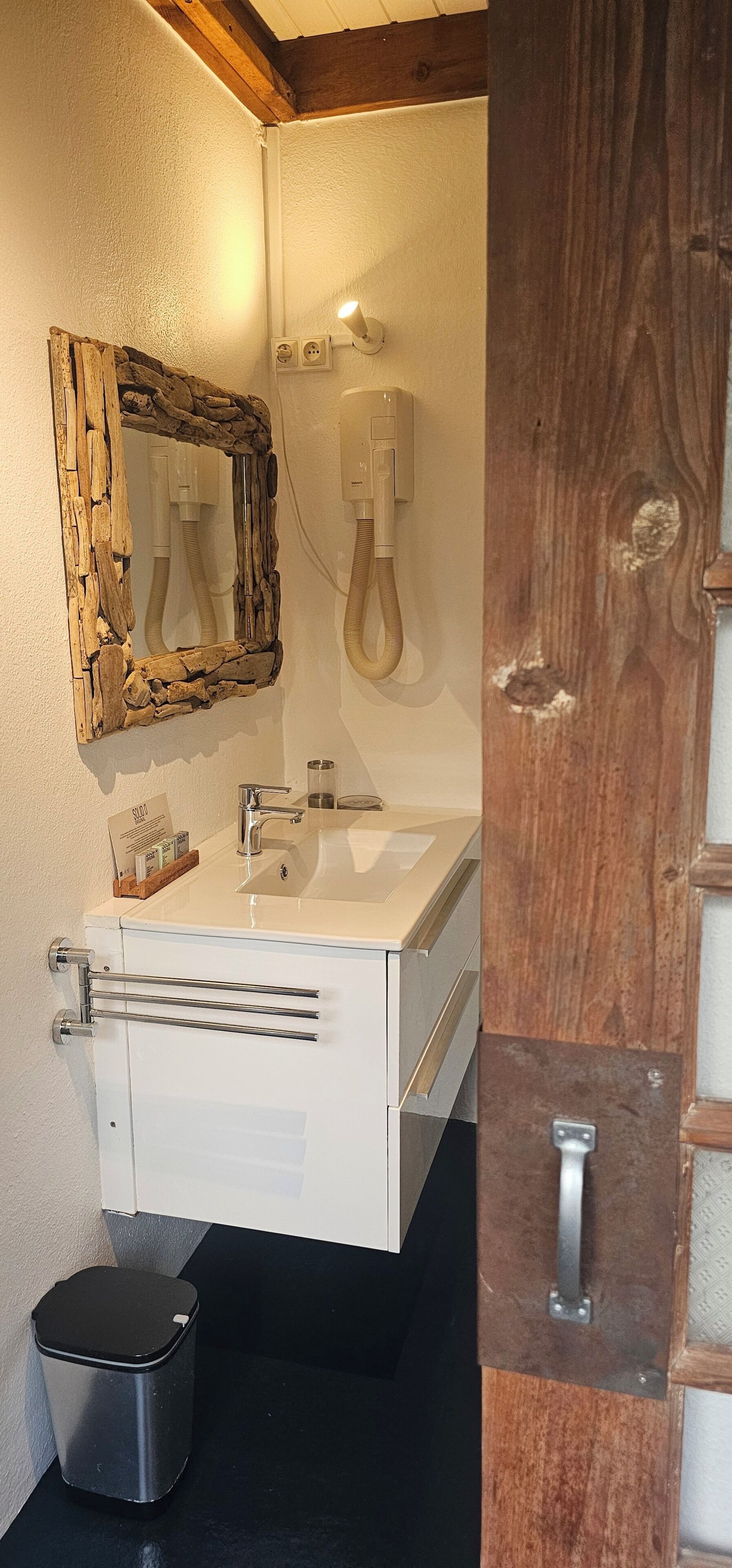 A bathroom with a white sink, a wooden-framed mirror, a wall-mounted light, and a trash bin.