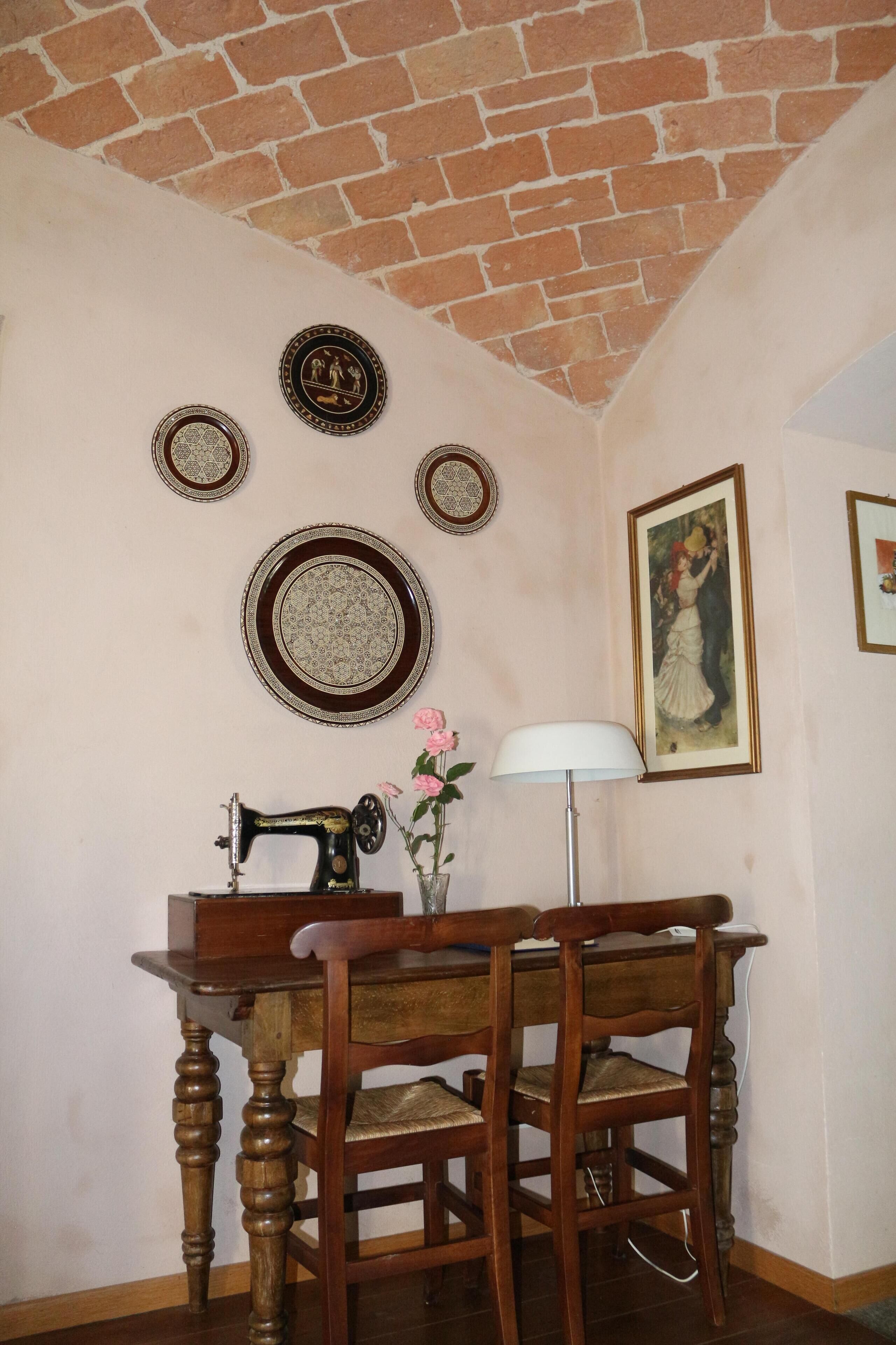 Une pièce avec un plafond en briques, une table en bois entourée de chaises, une machine à coudre, des assiettes décoratives et un tableau encadré.