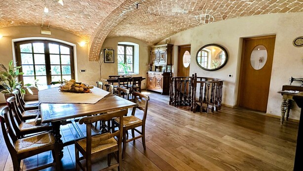 A spacious dining area with a large wooden table and chairs, a brick ceiling, and a view of the kitchen in the background. - Beautifully restored farmhouse with pool immersed in the greenery (MAGLIANO ALFIERI)