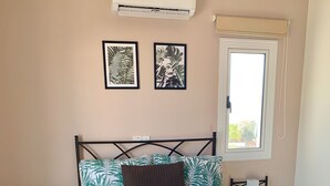A bed with a leaf-patterned bedspread, two pillows, and two rolled towels. There is a small bedside table with a glass top and a power outlet. A window with a view of buildings is visible. - Private Villa With Large Private Pool Situated In Coral Bay, Paphos, Cyrprus (coral bay)