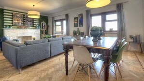 A spacious living area with a large sofa, dining table, and chairs. There is a fireplace, artwork on the walls, and large windows with curtains.