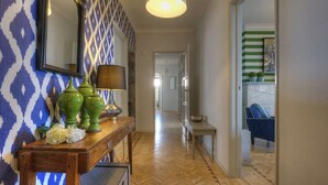 A hallway with patterned wallpaper, a wooden console table with decorative items, and a view into a room with a striped wall and a blue chair.
