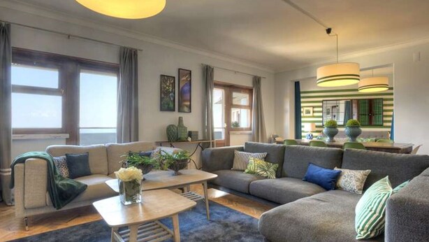A modern living room with a grey sofa, a coffee table, and a blue rug. There are large windows with curtains, a painting on the wall, and a hanging light fixture.
