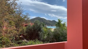 Paesaggio costiero con montagna, spiaggia e un edificio parzialmente nascosto dalla vegetazione.