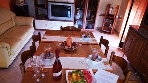 A dining area with a wooden table set for four, surrounded by chairs. A bottle of red wine and a fruit platter are on the table. - Villa Artemide with WI-FI 15 Km from Syracuse (siracusa)