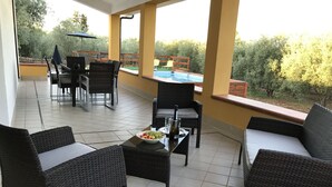 An outdoor patio with wicker furniture, a dining table, and a view of a swimming pool and greenery. - Villa Artemide with WI-FI 15 Km from Syracuse (siracusa)