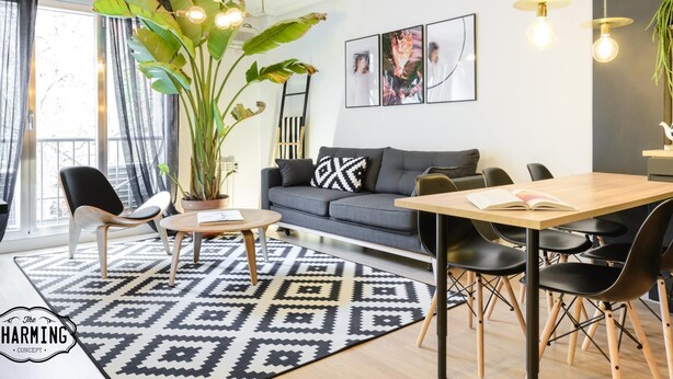 Ein modernes Wohnzimmer mit einer grauen Couch, einem schwarz-weißen Teppich, einem Couchtisch aus Holz und einem Essbereich mit einem Holztisch und schwarzen Stühlen. Eine große Pflanze in einem Topf und eine Hängeleuchte befinden sich über der Couch.